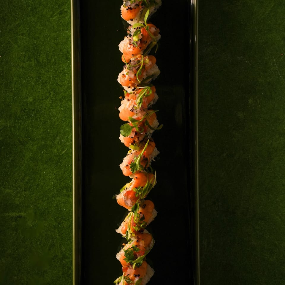 A long, dark plate holds a row of sushi rolls topped with orange sauce and green garnishes.