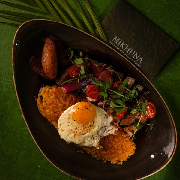 Mikhuna's Japanese-Peruvian dish with fried plantains, rice, egg, tomatoes, and meat on a dark plate.