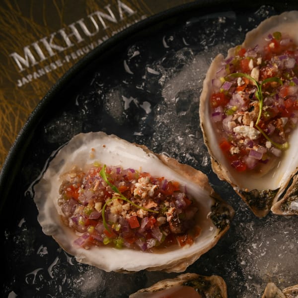 Two Mikhuna Japanese-Peruvian oysters on ice, topped with diced red onion, tomato, and green garnish.