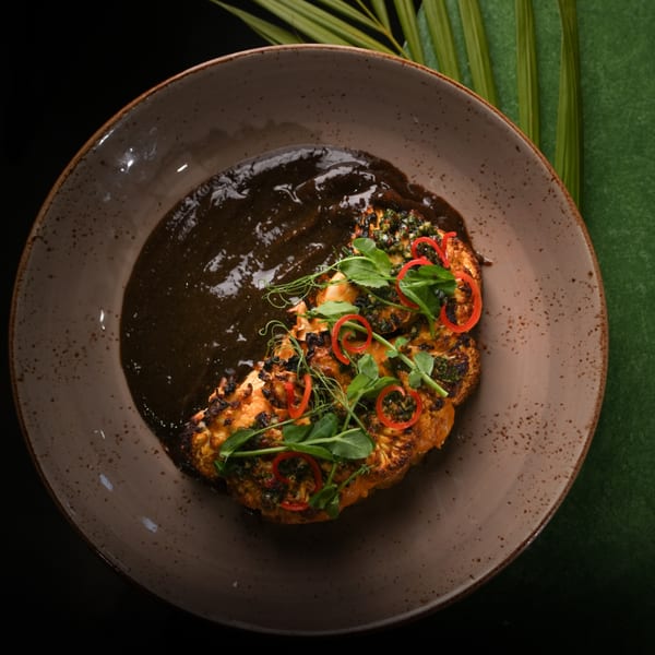Grilled cauliflower steak with dark sauce, red chili, and green herbs on a brown plate.