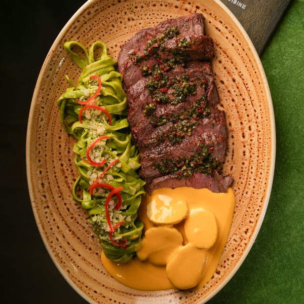 Mikhuna Japanese-Peruvian dish with sliced steak, green pasta, and yellow sauce on a speckled plate.